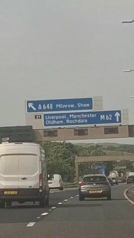 The video shows a highway sign indicating directions to A640 Milnrow, Shaw. Another sign below points to Liverpool, Manchester, Oldham, and Rochdale via M62. The camera is positioned inside a vehicle, showing the road ahead with other cars and trucks. Streetlights line the right side of the highway. The sky is overcast.