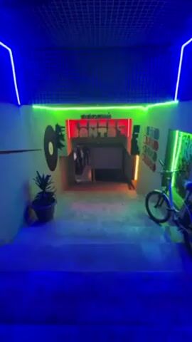 The video shows an entryway. The scene is lit with blue, green, and red neon lights. The walls are painted grey and feature painted silhouettes of a record, the Simpsons characters, and video game controls. There's also a plant in a pot and a bicycle visible. Through the entryway, people are standing in an indoor space with a screen.