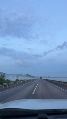 The video shows a view from inside a vehicle traveling on a highway. The sky is overcast with blue and gray clouds. There is a low-lying fog or mist along the sides of the road, obscuring the landscape. A truck with red taillights is visible in the distance on the highway. The road has white lane markings. The camera appears to be mounted on the dashboard or windshield of the vehicle.