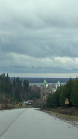 The video shows a road leading downhill towards a church in the distance. The church has green domes. On either side of the road are trees. A speed limit sign is on the right. The sky is overcast and gray.