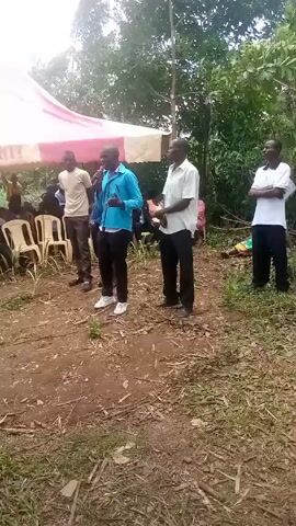 The video shows an outdoor gathering, possibly a memorial or similar event. Four men stand in a line facing the camera. The second man from the left speaks into a microphone while gesturing with his left hand. Behind them, under a white tent with a red stripe along the edge, people sit on white chairs. The ground is a mixture of dirt and patchy grass. Trees surround the area, suggesting a rural or semi-rural location.
