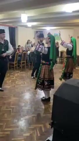 The video shows four individuals performing a traditional dance in a room with an audience. Two men and two women are dressed in colorful folk costumes, including black vests, trousers, and skirts, with accents of red, green, and white. The women wear green headscarves. The dancers move in unison, with their arms raised. They are standing on a patterned wooden floor. In the background, there are people sitting at tables, watching the performance. A speaker is partially visible in the foreground. Overhead lighting illuminates the room.