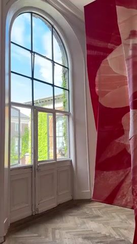 The video pans through a room with large, arching windows. The windows offer a view of ivy-covered walls and a blue sky. Inside the room, several large fabric banners hang from the ceiling. The banners are red and white and feature close-up images of flowers. The floor is made of parquet. The video shows multiple banners, some appearing in the foreground and others in the background. One banner in the background appears to be partially obscured by another. Towards the end of the video, a person is briefly visible in the background, walking away from the camera.