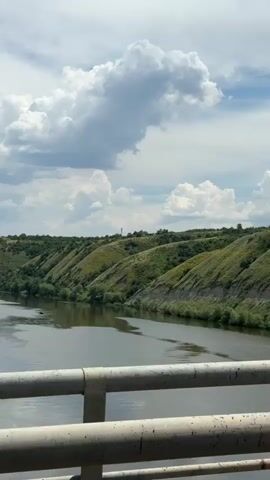 A vertical video shows a view from a bridge overlooking a wide river. On the opposite bank, rolling green hills covered in vegetation rise steeply from the water. The sky is partly cloudy with large white clouds. The railing of the bridge is visible in the foreground. The water is calm with some ripples. The video pans slowly to the right, revealing more of the river and the hilly landscape. There are no people or vehicles visible in the video, except for a small red object on the far right bank which is too indistinct to identify. The overall impression is one of a scenic, natural landscape.
