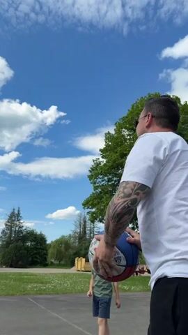 Here is a detailed description of the video:
The video shows a man and a boy playing basketball on an outdoor court on a sunny day. The sky is blue with scattered white clouds. The man is wearing a white t-shirt, black pants, and glasses. He has tattoos on his arm. The boy is wearing a green t-shirt, blue shorts, and a baseball cap. The man is shooting a basketball towards the hoop. The ball bounces off the rim and the boy catches it. The background includes a green lawn, trees, a small building, and houses in the distance. The camera is at eye level and follows the action as the man shoots and the boy retrieves the ball.