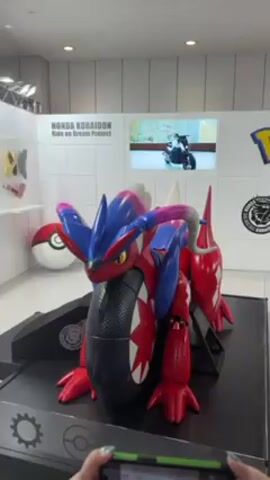 The video shows a display featuring Pokémon Koraidon and Miraidon statues, part of the "Honda Koraidon Ride on Dream Project" and "Toyota Miraidon Project." The Koraidon statue is predominantly red and blue, posed on a black platform with gear-shaped decorations. A Poké Ball is visible in the background. The camera pans to show the "Pokémon" logo on a white wall, accompanied by motorcycle-themed graphics. The Miraidon statue, primarily purple with neon accents, is positioned nearby on a similar platform. A small video display shows workers engaged in a related activity. The scene appears to be inside a large, well-lit exhibition space. Someone is recording the scene with a smartphone.