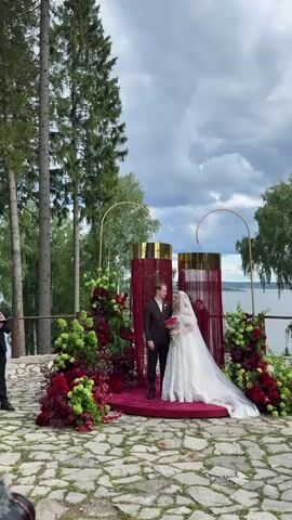 A couple, a man and a woman, dressed in wedding attire, stand on a circular red platform at an outdoor wedding ceremony. The woman wears a white wedding dress with a veil, and the man wears a dark suit. They are positioned in front of a wedding arch decorated with red fabric and gold accents, flanked by elaborate floral arrangements in shades of red and green. The ceremony takes place on a stone-paved area with tall trees in the background and a body of water visible in the distance under a cloudy sky.