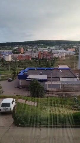 This is a short video clip taken from an elevated viewpoint, looking out over a town. The scene depicts a residential and commercial area with a mix of buildings, including apartment blocks and what appears to be a commercial establishment with a blue roof. In the foreground, there is a white car parked on a paved area, and a grassy slope with some wildflowers.

The overall setting is a town in Zheleznogorsk-Ilimsky, Russia. The time of day appears to be late afternoon or early evening, as the sky is overcast with diffused light, suggesting a cloudy day or approaching sunset. The weather seems mild, with the greenery indicating a period of growth.

There are no people clearly visible in the clip, but there are cars on the road, suggesting normal activity within the town. A dark-colored car is visible driving on the road near the blue-roofed building.

Notable details include the architecture of the buildings, with several multi-story apartment buildings visible in the distance, characterized by brick facades. The commercial building with the blue roof has a flat roof with some equipment on it. The foreground shows a slightly unkempt but natural area with vibrant green grass and scattered white flowers. The sun's rays are subtly visible as streaks in the lower right corner, suggesting it's trying to break through the clouds.

No visible text is present in the media file. The overall mood conveyed is one of everyday life in a moderately sized Russian town, under a somewhat subdued sky.