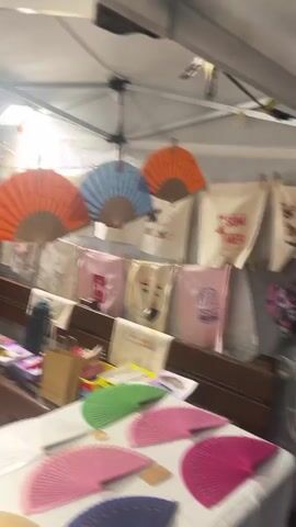 The video shows a market stall with various items for sale. A woman is sitting at a table, possibly attending the stall. The stall displays a variety of colorful fans, both open and closed, as well as fabric bags with designs. There are also racks of jewelry, including necklaces and earrings, and a display of bracelets. The market appears to be outdoors or in a covered area, possibly a tent, with overhead lighting. The atmosphere seems to be that of a casual outdoor market or craft fair.