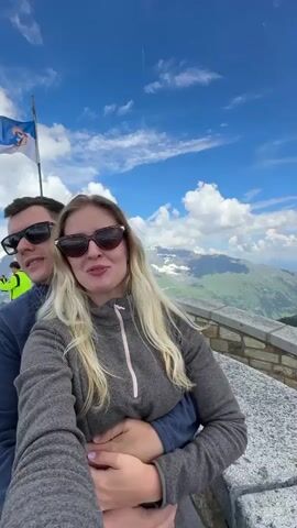 A couple is taking a selfie on a mountain with a scenic view. The man is wearing a dark blue sweater and sunglasses, and he has his arm around the woman. The woman is wearing a grey fleece jacket and sunglasses, and she has long blonde hair. They are both smiling at the camera. In the background, there are mountains with snow on top and a bright blue sky with white clouds. There are other people in the background, as well as a stone railing and a sign. The overall scene is a beautiful, clear day in the mountains.