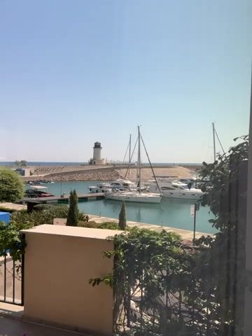 A daytime, outdoor, medium shot captures a bright, sunny scene of a marina. The view is from a higher vantage point, looking down and across the water. In the foreground, a tan-colored wall is visible, partially obscured by lush green foliage. Beyond the wall, a black metal fence runs horizontally across the bottom of the frame. The marina itself is filled with several white yachts and sailboats docked in the clear, turquoise water. Tall masts rise from the sailboats. In the background, a breakwater made of large rocks extends into the sea, and a white lighthouse with a black top stands on the end of it. The sea stretches out to the horizon under a clear, pale blue sky. The overall impression is one of a peaceful and picturesque coastal setting.