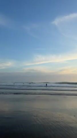 The video shows a beach scene at sunset. The sky is a mix of blue, white, and orange. There are a few people visible in the water, presumably surfing. The waves are small and gentle. The sand is wet and reflective, mirroring the sky. The sun is setting behind a small hill or cliff covered with trees.