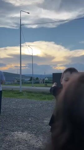 This is a video of a group of people outdoors. In the foreground, a young boy with dark hair is looking at the camera and smiling. He is wearing a black t-shirt. Behind him, a man in a grey t-shirt and dark pants is standing. To his left, a woman in a light-colored shirt is visible. Further in the background, there are buildings, streetlights, a road with a car, and a body of water. The sky is cloudy with the sun breaking through.