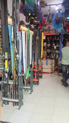A medium shot captures the interior of a fishing supply store. The camera pans slowly from left to right, showcasing rows of fishing rods stored in vertical racks along the left wall. These rods vary in color and length, with some in protective cases. Above the rods, fishing nets and life vests are displayed. The center of the store features a checkout counter where a person wearing a light green t-shirt and dark pants is interacting with a customer. Behind the counter, shelves are stocked with various fishing gear, including spools of fishing line and tackle boxes. To the right, a glass display case showcases more fishing supplies. The store's walls are lined with shelves filled with merchandise, and the floor is tiled. The lighting is bright, illuminating the store's offerings.