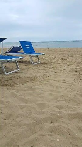 This video shows a beach with several empty lounge chairs and a few beach chairs. The sand is light brown and the water is a calm blue. The sky is overcast. In the distance, a lighthouse is visible. There are no people in the video. The video appears to have been taken from a low angle, with the camera pointed towards the ocean. The lounge chairs are blue and white, and the beach chairs are blue and white. The lounge chairs are arranged in rows, and the beach chairs are scattered around. The overall impression of the video is one of a peaceful and relaxing beach.
