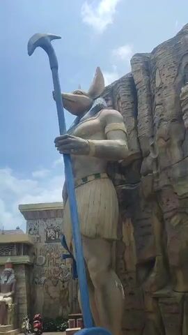 The video pans across an Egyptian themed area. On the right, there is a large statue of Anubis, the Egyptian god of the dead, holding a blue staff. In the background is a building with carvings depicting Egyptian scenes and hieroglyphs. There is a seated statue of a pharaoh in front of the entrance. The sky is blue with some clouds. There is a scooter parked near the building. The building is labeled "The Temple of Isis".