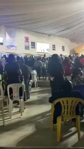 The video depicts a large group of people gathered inside a spacious, brightly lit venue with a tented ceiling. Many are standing, some seated on white plastic chairs, and they appear to be interacting with each other and the stage area. The crowd is diverse in terms of clothing and age. There are multiple screens displaying images or videos, and decorative banners hang in the background. The atmosphere seems lively and possibly celebratory.