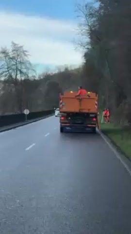 The video shows an orange dump truck stopped on the right side of a road, adjacent to a grassy shoulder. There's a person in an orange safety suit sitting on top of the truck bed. Two other people, also wearing orange safety suits, are standing next to the truck on the grassy shoulder, one holding a long pole with reflective markings. Cars are driving on the road, including a white Tesla. The environment includes trees and foliage on the side of the road, and the sky is overcast with blue patches. The road is wet, suggesting recent or ongoing precipitation.