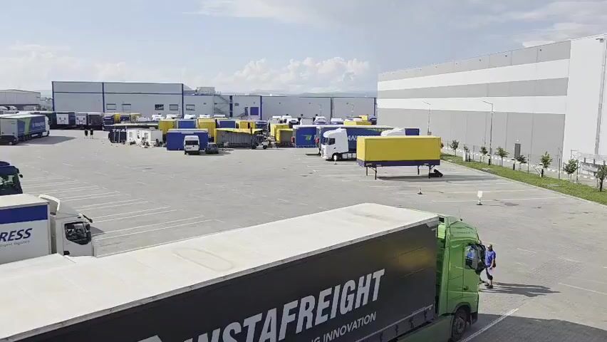 This video provides an aerial view of a large, active logistics and warehouse complex, likely located in an industrial zone like Bozhurishte, Bulgaria. The scene is set on a bright, clear to partly cloudy day.

The primary subjects are numerous semi-trailer trucks and their trailers, occupying a vast paved parking lot. In the foreground, a prominent green truck with a black trailer clearly displays the company name "INSTAFREIGHT" with "INNOVATION" written below it. A person, dressed in blue, walks from the right side of the frame towards the left, near this green truck. On the far left, a white truck is partially visible, bearing the text "XPRESS."

Throughout the extensive parking area, various trucks and trailers are parked. Many trailers are distinctively colored in bright yellow or blue, while others are plain white or grey. Several smaller blue container-like structures are also scattered in the central part of the lot, possibly serving as temporary offices or storage units.

The perimeter of the parking lot is defined by large, modern industrial or warehouse buildings, characterized by their light grey and white horizontally striped facades. Along the right side of the main building, a strip of green landscaping with small trees and a fence adds a touch of greenery to the otherwise industrial setting. The overall activity suggests a typical operational day at a busy logistics hub, with vehicles parked, waiting, or potentially being prepared for loading and unloading.
