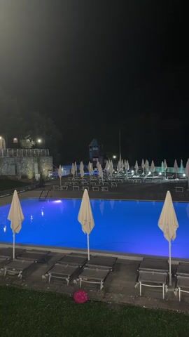 The video shows a nighttime view of an empty swimming pool. The pool is illuminated with blue lights, casting a glow on the water. Deck chairs and closed umbrellas are arranged around the pool area. In the background, there are trees and some structures, possibly part of a resort or park. The overall atmosphere is calm and still, with no people visible.