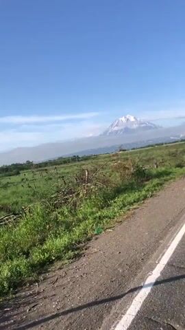 The video shows a moving camera perspective, likely from a vehicle, traveling along a paved road. The road has a white line marking its edge. To the left of the road is a grassy field with some bushes and trees. In the distance, a large mountain, possibly a volcano, is visible, capped with snow and surrounded by clouds. A triangular road sign is also present on the left side of the road, mounted on a pole. The sky is blue and clear. The sun appears to be shining, casting shadows on the road. The overall scene is a rural landscape with a prominent natural feature.