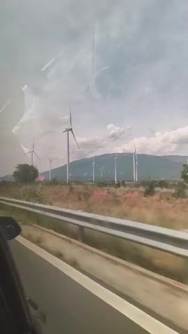 This video shows a view from a moving vehicle looking out at a field of wind turbines on a cloudy day. The turbines are tall and white, with rotating blades. In the foreground, there is a road or highway with a guardrail and some greenery. The background consists of rolling hills covered with trees and vegetation. The sky is overcast with dramatic cloud formations. There are no people visible in the video. The overall scene suggests a rural or semi-rural landscape with renewable energy infrastructure.