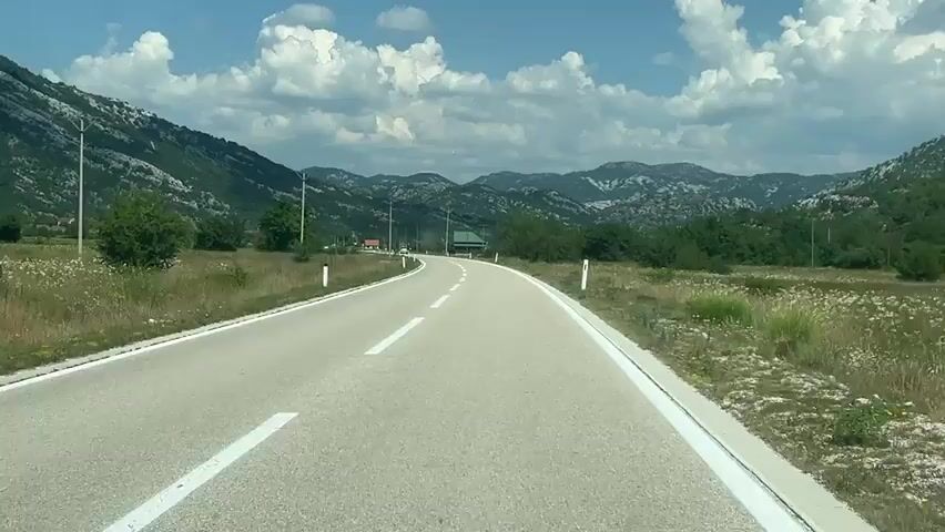 The video shows a view from a vehicle driving on a paved road through a mountainous landscape. The road curves to the left, with white lines marking the lanes. On either side of the road, there are grassy fields and scattered trees. In the distance, there are more hills and mountains under a blue sky with fluffy white clouds. There are no people visible in the video. The overall environment is rural and scenic.