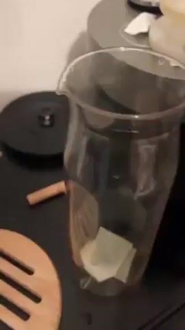 Someone pours hot water into a clear glass pitcher, which already contains green herbs and water. The pitcher is on a black surface with a cinnamon stick and a wooden trivet next to it. A coffee maker is slightly visible in the background.