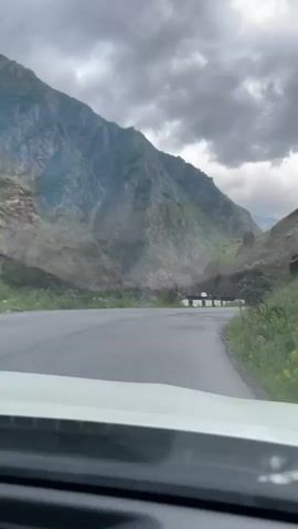 The video shows a view from the front seat of a car driving on a winding mountain road. The road surface is asphalt and appears weathered with some potholes. On either side of the road are steep, grassy mountainsides. The sky is overcast with dramatic, dark clouds. In the distance, a white vehicle is visible on the road ahead, and in some frames, a guardrail is visible along the edge of the road. The mountains are rocky and appear to have areas of loose scree and vegetation. The overall environment suggests a dramatic and somewhat remote mountainous landscape.