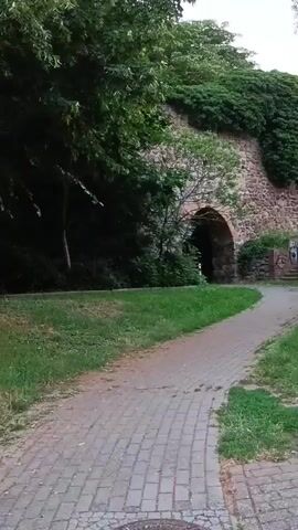 The video pans across a cobblestone path bordered by grass and trees. In the background, an ancient stone wall with an arched entrance is visible. Further along, the path leads past modern apartment buildings with red tiled roofs. A person is seen walking on the path in the distance.