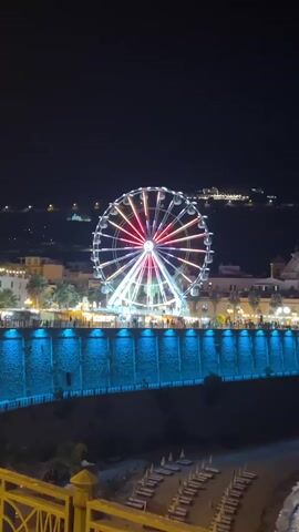 The video shows a Ferris wheel lit up with colorful lights at night. The Ferris wheel is located in a town or city with buildings and lights visible in the background. There is a wall with blue lights along its base in the foreground. The bottom of the video shows a beach with lounge chairs. The Ferris wheel appears to be stationary.