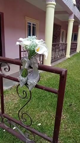 A pair of silver, embellished high-heeled shoes and a bouquet of white calla lilies and baby's breath are hanging from a dark metal railing. The railing is in front of a green grassy area. Behind the grass is a light pink building with white columns and windows. The lighting suggests it is daytime.