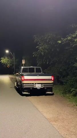 A dark-colored pickup truck with chrome trim and dual exhaust pipes is parked on the side of a road at night. The truck's brake lights are illuminated, and the license plate is visible. Trees and foliage are present on the right side of the road, and a street light illuminates the left side. The truck appears to be a Ford F-Series, likely from the late 1980s or early 1990s. The scene is dimly lit, suggesting it is nighttime.
