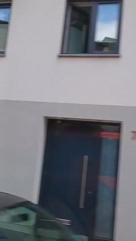 The video shows a view from inside a vehicle, possibly a bus or car, as it travels through a residential area. The camera pans from right to left, capturing a series of houses and buildings. 

In the first few seconds, a white car is visible parked in a driveway, partially obscured by a structure that appears to be a carport or garage. The car has a yellow sign behind it. 

The view then shifts to the side of buildings with white facades and green window frames. Further along, a house with a wooden balcony with planters is seen, with a white garage door below it. Another building with white stucco and light blue window frames appears, with a white car parked in front of it. 

The video continues to show more residential buildings with various architectural styles, including some with timber-framed elements and dark tiled roofs. Several windows are visible, some with curtains. Parking signs are also observed. The sky is overcast.