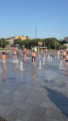 The video shows a group of children playing in a public fountain on a sunny day. The fountain consists of multiple water jets spraying upwards from the ground. Several children are seen running through the water jets, splashing and laughing. One child in the foreground is doing a dance-like movement while jumping over a water jet. In the background, there are buildings, trees, and people walking. The ground around the fountain is paved with gray tiles. The overall atmosphere is one of playful enjoyment.