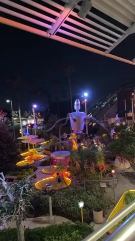 The video shows an elevated view of a miniature golf course at night. A large robot figure stands at the center of the course, and various obstacles are illuminated with colored lights.  The course appears to be Bubba Gump Shrimp Co. themed. The video then transitions to an escalator within a covered outdoor mall-like area.  Signs are overhead, including directions to Universal Studios Florida.  People are walking on the escalator and throughout the area.