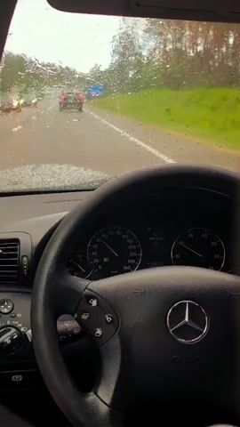 The video shows a first-person view from inside a Mercedes-Benz car driving on a wet road during rainfall. The windshield wipers are seen clearing the rain. The dashboard instruments, including the speedometer, are visible. A hand is operating the turn signal stalk. Other cars are visible on the road ahead. The environment appears to be a rural or suburban area with trees and grass alongside the road.