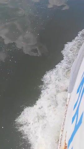 This is a close-up, high-angle shot of water and the side of a boat. The water is a dark gray-green color and appears choppy with white foam trailing behind the boat. Reflections of clouds are visible on the surface of the water. The side of the boat is white with large blue letters printed vertically on it. The letters appear to spell out "AZUL". The video is taken from the perspective of someone on the boat looking down and to the side, as the boat moves forward through the water.