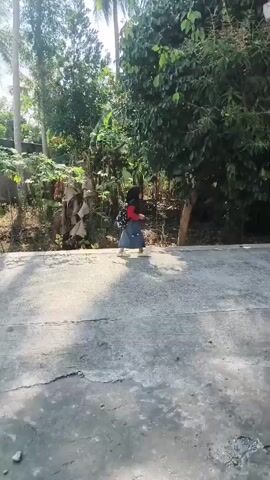 A child wearing a red hat and blue shorts is walking down a concrete path. The path is cracked and worn. To the left of the path, there are trees and foliage. To the right of the path, there is a building with a white roof and more trees and foliage. The sun is shining brightly, casting shadows on the path. The child is walking away from the camera.