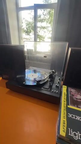 A close-up, eye-level shot shows a record player on an orange surface, with a vinyl record spinning on it. Two dark speakers are visible on either side of the record player, and a stack of records is on the right. Sunlight streams through a window in the background, illuminating the scene. The overall mood is nostalgic and warm.