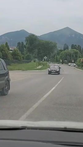 The video shows a view from inside a vehicle looking out at a road with mountains in the background. The sky is overcast. Several cars are visible on the road. The vehicle appears to be moving forward. There is greenery and trees along the side of the road, as well as some buildings. Traffic signs are also visible.