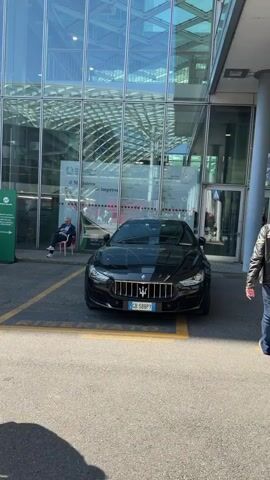 The video captures scenes outside and within a modern architectural complex, likely the Fiera Milano exhibition center in Rho, Italy, on a clear, sunny day. It begins with a black Maserati Ghibli and a black Maserati Grecale/Levante SUV parked in front of a glass building, where a person is seated next to a green sign. Further along the building, a large banner displays "MILANO IS MORE" alongside Olympic rings and Paralympic symbols. People are seen walking along a landscaped pathway and under a prominent glass and metal canopy structure. A red pop-up store with "POP UP STORE AND GOODS" written on it is also visible amidst the crowd. Italian license plates "CB 588PY" and "CE 812XX" are seen on the Maserati vehicles.