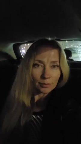 A medium close-up video shows a blonde woman sitting in the back seat of a car at night. She is looking directly at the camera and smiling faintly. Her long, light-colored hair falls over her shoulders. She is wearing a dark top with a striped pattern visible at the neckline. The interior of the car is dark, with some light illuminating her face, likely from the screen she's recording with or ambient streetlights. Through the rear window, blurred lights and streaks of rain are visible, suggesting it's raining and the vehicle is in motion. The overall atmosphere is calm. No visible text is present. The scene is set in Vecsés, Hungary.