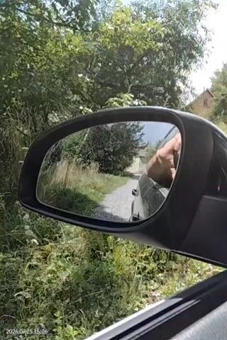 The video is taken from inside a car, looking out the passenger side mirror. The mirror reflects a dirt road or path lined with tall grass and trees. A person's hand is visible in the reflection, appearing to be holding onto the side of the car or leaning out the window. In the background, there are houses with roofs visible through the trees. The timestamp in the lower left corner reads "2026.07.25 15:26".