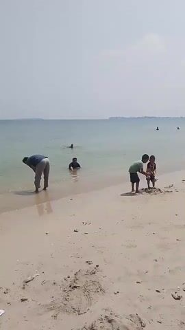 A wide shot of a sandy beach and calm ocean under a hazy sky. In the foreground, the sand is littered with debris. Further back, near the water's edge, a person in a dark shirt and light pants is bent over, appearing to pick something up from the shallow water. To their right, two young boys are playing in the sand, one of them squatting while the other stands. In the water further out, two other individuals are visible, one swimming and another standing waist-deep. In the distance, the faint outline of land is visible across the water.