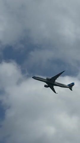 A commercial airplane is filmed from below as it flies through a partly cloudy sky. The plane is white with blue accents on its tail and appears to be a wide-body jet. It is captured in flight from various angles as the camera pans and zooms.