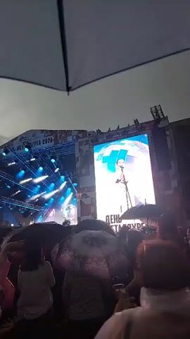 The media file shows an outdoor event with a large stage set up. A big screen behind the stage displays a person singing, with "ДЕНЬ " visible at the bottom of the screen. The stage is lit with numerous blue lights. Text reading "МЕТАЛЛУРГА 2026" is also visible on the stage structure. A crowd of people, many holding umbrellas, are gathered in front of the stage, suggesting it is raining or has recently rained. The sky appears overcast, indicating either early morning or late afternoon, or simply cloudy weather. The atmosphere seems to be festive, with some individuals appearing to sing along or react to the performance. The presence of umbrellas and the mood of the crowd point towards a potentially challenging but enjoyable experience for attendees at this outdoor festival in Solnechnyy, Russia.