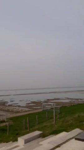 The video shows a landscape view from an elevated position, likely a set of stairs or a low hill. The foreground includes concrete steps and a patch of green grass fenced with wooden posts. Beyond the grass, there's a tidal area with mudflats and patches of water. A rocky pathway stretches out into the water. In the distance, there is a pier with people on it. The sky is overcast and gray. The camera pans to the left, revealing a trash can and a signpost, before panning back to the right.