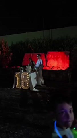 A person is standing behind a table with a white tablecloth. There is a cake on the table and large light-up numbers that spell out "18". Behind the table there is a red-lit rock formation. To the left of the table, another person is standing. Both people appear to be looking at the cake.