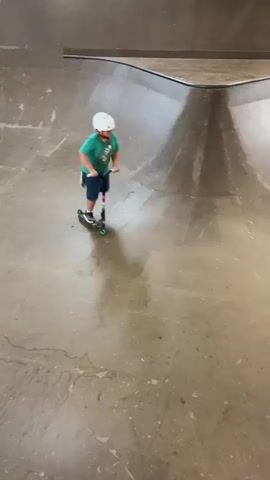 A child wearing a green shirt, blue shorts, a white helmet, and white shoes is riding a scooter in a skate park. The child moves from left to right across the frame, then turns and rides up a curved wall. The child then falls off the scooter and lands on their back, the scooter continues moving up the wall and falls to the ground. The child then pushes themselves up and slides down the wall. The skate park appears to be made of wood with concrete accents.